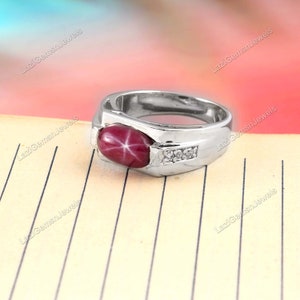 Può includere: Un anello d'argento con una grande gemma di rubino a stella ovale. Il rubino è di colore rosso intenso con un motivo a stella a sei raggi. L'anello presenta anche tre piccole pietre di accento trasparenti.