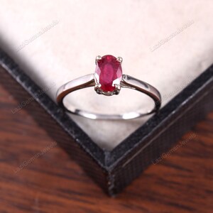May include: A silver ring with a red oval ruby gemstone. The ring is in a black ring box.