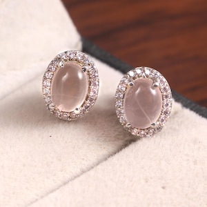 Op de afbeelding: Een paar ovale oorstekers. Elke oorbel heeft een lichtroze edelsteen omgeven door een halo van kleinere, heldere en roze stenen. De oorbellen zijn gezet in een zilverkleurig metaal en worden gepresenteerd op een lichtgekleurd oppervlak.