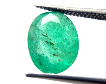 Pode incluir: Uma gema de esmeralda verde vibrante com forma oval facetada, segurada por pinças sobre um fundo branco.