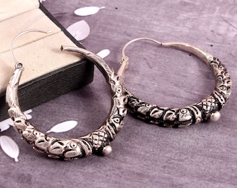 May include: A pair of silver hoop earrings with intricate, detailed designs. The earrings are circular and have a textured, ornate finish.