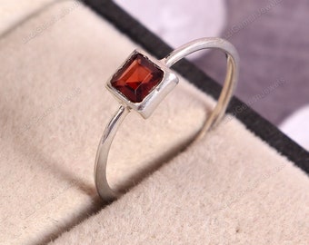 May include: A silver ring featuring a square-cut, deep red gemstone. The gemstone is set in a square bezel, and the band is thin and polished. The ring is displayed on a neutral-toned surface, highlighting the jewelry's design.