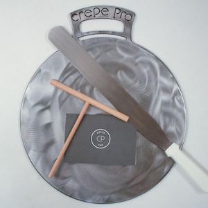 May include: A stainless steel crepe griddle with a handle, a long spatula with a white handle, and a wooden crepe spreader. A dark gray card with the text "Crepe Pro" and a logo is also included.