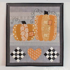 May include: A patchwork quilt featuring two orange pumpkins with different patterns, a heart, and two black and white checkered squares. The quilt is framed with a black and white checkered border. The text "Patchwork Pumpkins" is at the top of the image and "the crafty quilter" is at the bottom.