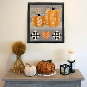 May include: A grey wooden console table with two drawers features a fall-themed display. A bunch of dried wheat stalks, a white knitted pumpkin, a mosaic pumpkin, and a black candle holder with a white candle are arranged on the table. A framed quilted wall hanging with two orange pumpkins and a heart is displayed on the wall above the table.