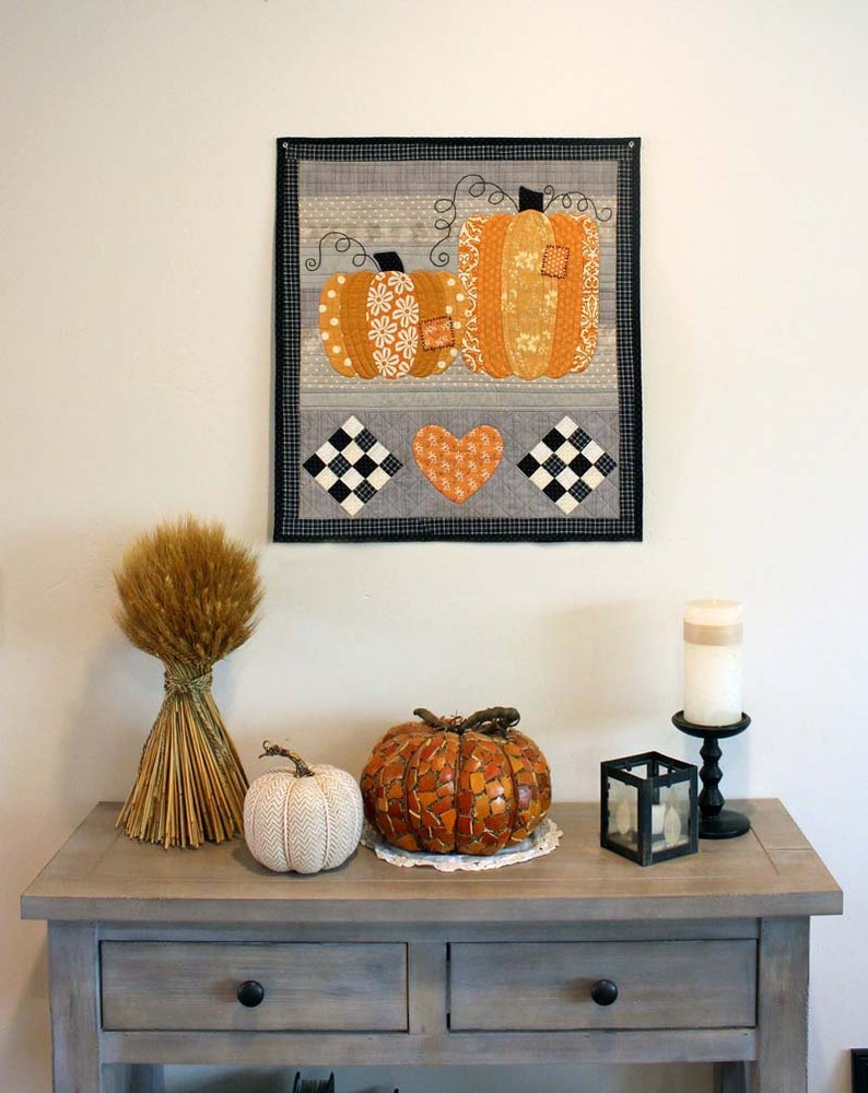 May include: A grey wooden console table with two drawers features a fall-themed display. A bunch of dried wheat stalks, a white knitted pumpkin, a mosaic pumpkin, and a black candle holder with a white candle are arranged on the table. A framed quilted wall hanging with two orange pumpkins and a heart is displayed on the wall above the table.