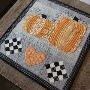 May include: A grey and black quilted table runner with a patchwork design featuring two orange pumpkins and a heart. The pumpkins have a white floral pattern and the heart has a swirl design.