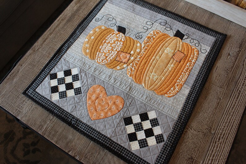 May include: A grey and black quilted table runner with a patchwork design featuring two orange pumpkins and a heart. The pumpkins have a white floral pattern and the heart has a swirl design.