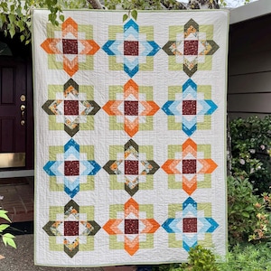 May include: A handmade quilt with a repeating geometric pattern. The design features squares in shades of orange, blue, green, and brown, set against a white background. The quilt is hanging outdoors, showcasing its colors and craftsmanship.