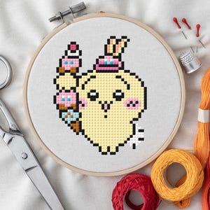 Könnte beinhalten: Ein Kreuzstich-Stickrahmen mit einem gelben, pixeligen Charakter, der Eiswaffeln hält. Das Design umfasst rosa, blaue und braune Details. Schere, Faden und Nadeln sind um den Rahmen angeordnet.