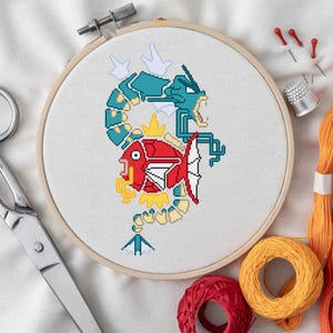 Könnte beinhalten: Ein Kreuzstich-Stickrahmen mit einem rot-blauen, pixeligen Design eines Fisches und eines Drachen. Das Bild enthält eine Schere, orangefarbenes und rotes Stickgarn sowie andere Nähutensilien.