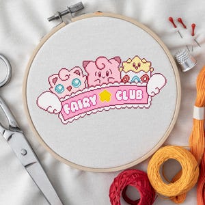 Könnte beinhalten: Ein Stickrahmen mit einem Kreuzstich-Design mit Cartoon-Figuren und dem Text "FAIRY CLUB" in Rosa. Das Bild enthält eine Schere, orangefarbenes und rotes Stickgarn, Nadeln und einen Fingerhut.