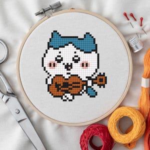 Könnte beinhalten: Ein Kreuzstich-Stickrahmen mit einer pixeligen Katze, die Ukulele spielt. Die Katze hat einen weißen Körper, blaue Haare und rosa Wangen. Schere, Faden, Nadeln und ein Fingerhut sind um den Rahmen angeordnet.