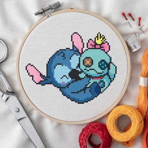 Pode incluir: Um bastidor de bordado em ponto cruz com uma imagem pixelizada de duas personagens de desenhos animados em tons de azul e verde-azulado, com detalhes em rosa. Tesouras, linha, agulhas e uma dedeira são visíveis.
