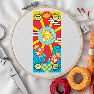 Könnte beinhalten: Kreuzstich-Stickrahmen mit einem farbenfrohen Design mit drei Fischen, Sonnenstrahlen und chinesischen Schriftzeichen. Die Stickerei befindet sich auf einem weißen Stoff, mit Schere, Faden und Nadeln in der Nähe.