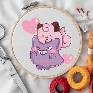 Könnte beinhalten: Ein Kreuzstich-Stickrahmen mit einem rosa-lila Cartoon-Charakter-Design mit Herzen. Das Bastelprojekt ist von einer Schere, Fäden, Nadeln und einem Fingerhut umgeben. Das Design befindet sich auf einem weißen Stoff.