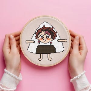 Puede incluir: Un bastidor de bordado de punto de cruz con un personaje de dibujos animados dise&ntilde;ado como una bola de arroz, o onigiri. El personaje tiene cabello casta&ntilde;o, una cara sonriente y lleva una falda marr&oacute;n oscuro. El bordado est&aacute; sobre tela beige.