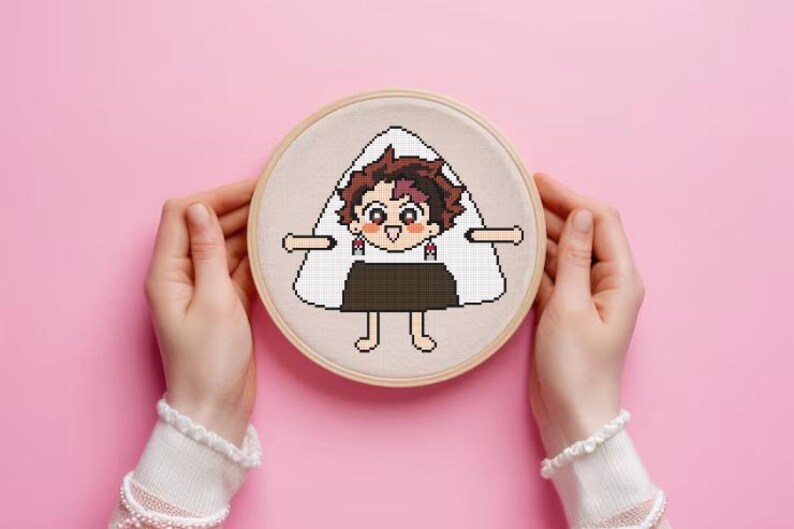 Puede incluir: Un bastidor de bordado de punto de cruz con un personaje de dibujos animados dise&ntilde;ado como una bola de arroz, o onigiri. El personaje tiene cabello casta&ntilde;o, una cara sonriente y lleva una falda marr&oacute;n oscuro. El bordado est&aacute; sobre tela beige.