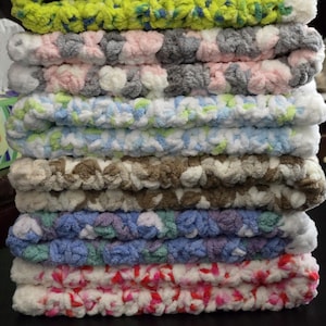 May include: A stack of crocheted blankets in various colors, including pink, blue, green, white, and brown. The blankets are made with a fluffy yarn and have a soft, textured appearance.