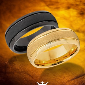 Op de afbeelding: Twee wolfraamringen, één zwart en één goudkleurig, worden getoond tegen een gouden achtergrond. De zwarte ring heeft een matte afwerking met een enkele groef, terwijl de gouden ring een getextureerde middenband heeft. De ringen zijn van Tungsten King.