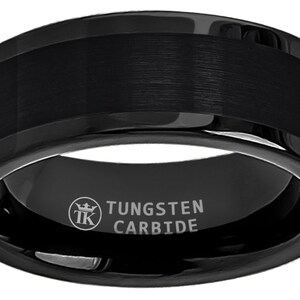 Puede incluir: Un anillo de carburo de tungsteno negro con acabado cepillado y borde biselado. El anillo está grabado con "TUNGSTEN CARBIDE" y un logotipo.