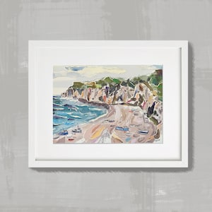 May include: A framed watercolour painting of a coastal scene. The artwork depicts a beach with small boats, cliffs, and ocean waves. The colour palette includes blues, greens, and earth tones. The painting is set in a white frame.