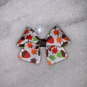 May include: A small, decorative bow featuring a white background with a repeating pattern of fall leaves and acorns in orange, brown, and green. The bow is tied with a brown center knot.