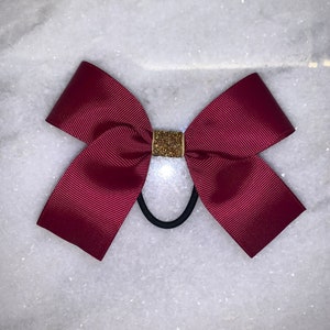 May include: A burgundy hair bow with a gold glitter center. The bow is attached to a black elastic hair tie.