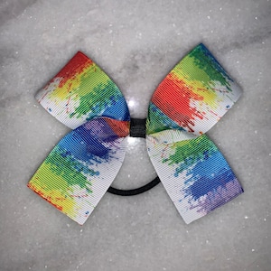May include: A white hair bow with a rainbow paint splatter design. The bow is tied with a black elastic band.