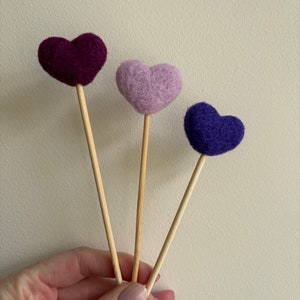May include: Three felt heart-shaped ornaments on wooden sticks. The hearts are purple, pink, and blue.