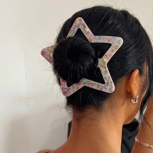 May include: A star-shaped hair clip with iridescent rhinestones is worn in the hair. The hair is styled in a bun. The clip is white with a clear outline. The person is wearing a black spaghetti strap top and silver jewelry.