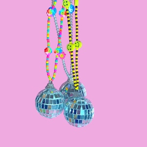 May include: Three disco ball ornaments with beaded strands in rainbow, silver, and black and yellow. The strands are adorned with smiley face and flower charms. The background is a solid pink.