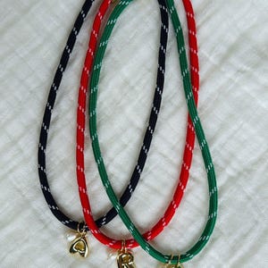 Peut inclure: Trois colliers en corde colorés, noir, rouge et vert, avec des fermoirs dorés et des breloques initiales. Les colliers ont des accents blancs. Chaque collier a une breloque initiale différente : A, R et C, avec une petite perle.