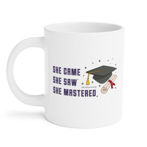 May include: White ceramic coffee mug with a graduation cap and diploma illustration. The text on the mug reads "She came, she saw, she mastered."  The text "@academemology" is also included on the mug.