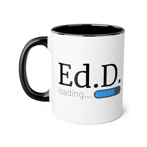 May include: White ceramic coffee mug with a black rim and handle. The mug has black text that reads "Ed.D. loading..." with a blue progress bar.