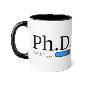 May include: White ceramic mug with a black rim and handle. The mug has a black graphic that says "Ph.D. loading..." with a blue progress bar.