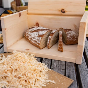 May include: A wooden bread bin with a hinged lid, open to reveal a loaf of sliced bread inside. The bread bin is made of light wood and has a natural finish. There is a pile of wood shavings in the foreground.