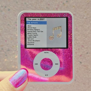 May include: A pink glitter iPod with a white screen displaying the text "The year is 2007" and a list of top artists from that year. The screen also shows a musical note icon. The iPod has a white control panel with buttons for play, pause, and skip.