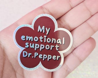 May include: A flower-shaped pin with a maroon center and a silver glitter outline. The pin reads "My emotional support Dr. Pepper" in white text. The pin is held in a hand.