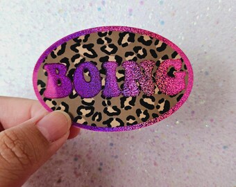 May include: A pink and purple iridescent sticker with a leopard print background and the word "BOING" in a retro font.