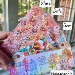 May include: A set of four decorative bookmarks with different designs. The bookmarks are made of clear plastic with a holographic finish. The designs include stars, dots, gems, and a house with trees. The quote "I am part of everything that I have ever read - Theodore Roosevelt" is printed on the house bookmark.