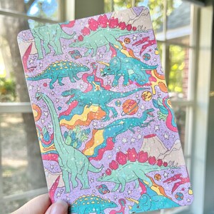 May include: A purple background with a repeating pattern of dinosaurs and planets. The dinosaurs are green, blue, and pink with glitter accents. The planets are blue, green, and orange. The background is covered in a rainbow pattern.