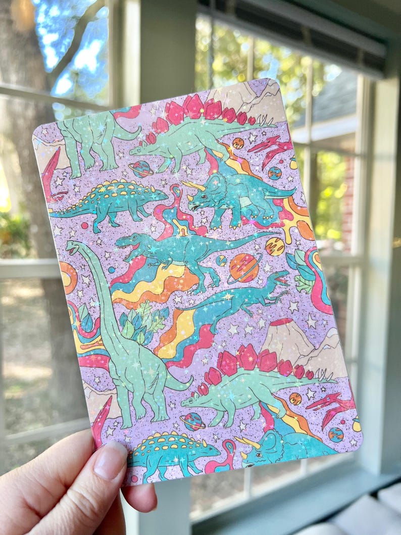 May include: A purple background with a repeating pattern of dinosaurs and planets. The dinosaurs are green, blue, and pink with glitter accents. The planets are blue, green, and orange. The background is covered in a rainbow pattern.