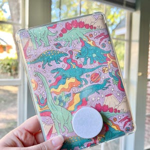May include: A clear phone case with a colorful dinosaur print. The case has a white pop socket attached to the back.