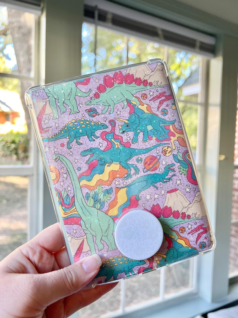 May include: A clear phone case with a colorful dinosaur print. The case has a white pop socket attached to the back.