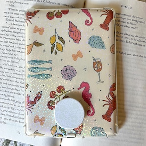 May include: A clear phone case with a whimsical design featuring sea creatures, lemons, and cherries. The case has a white pop socket with a floral pattern. The background is a book with text.