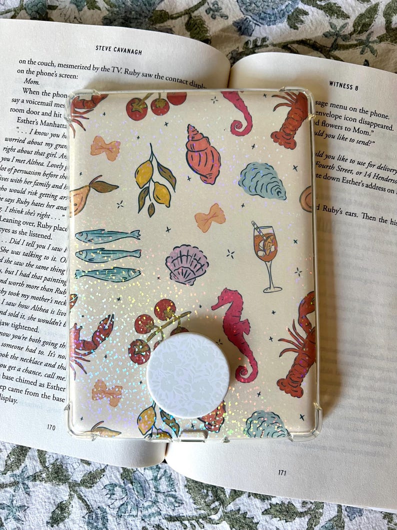 Pu&ograve; includere: Una custodia trasparente per telefono con un design stravagante con creature marine, limoni e ciliegie. La custodia ha un pop socket bianco con un motivo floreale. Lo sfondo &egrave; un libro con testo.