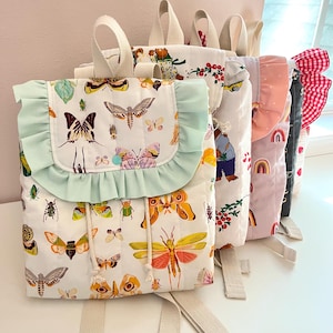 May include: A row of colorful, patterned backpacks. The first backpack features a white background with insect illustrations and a light green ruffled flap. Other backpacks have various patterns, including bears, rainbows, and cherries.