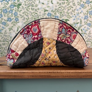 May include: A half-circle quilted pouch with a black zipper. The patchwork design features floral patterns in red, blue, and yellow, with beige and black sections. The pouch is on a wooden surface with a floral wallpaper background.