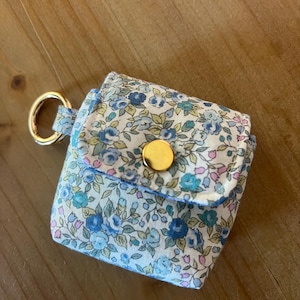 May include: A small, square fabric pouch featuring a light blue, teal, and white floral pattern. The pouch has a fold-over flap secured with a gold-colored metal snap button and includes a gold-colored metal ring attached to the side for hanging.